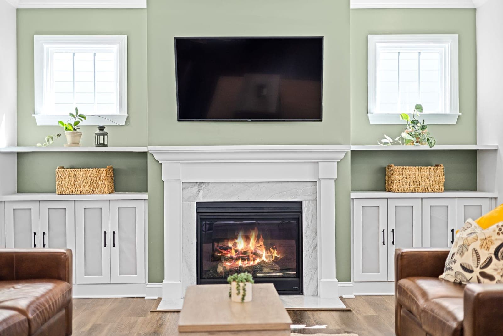 TV mounted above a white mantel with a lit fireplace and built-in cabinets on a sage green wall