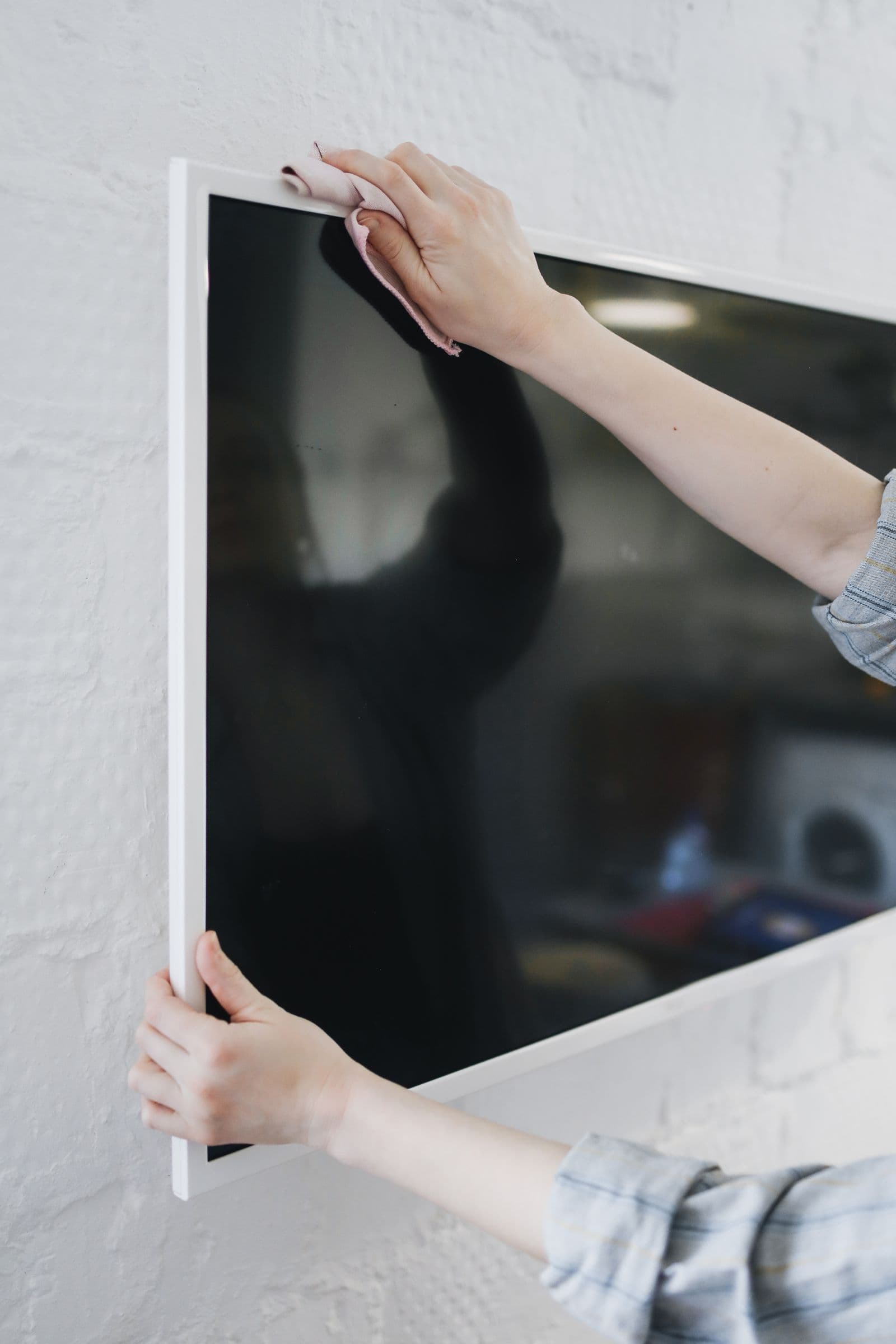 Hands wiping down a white-bezel TV mounted on a whitewashed brick wall